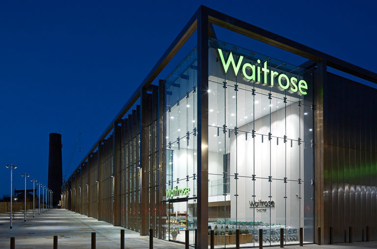 Evening photo of exterior façade to Waitrose in Chester, with full height glazing and bronze metal cladding elements.