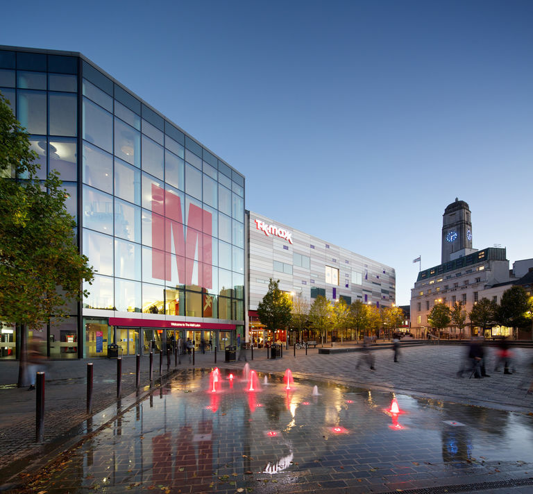 Evening photo of The Mall in Luton, showcasing a triple height fully glazed landmark entrance and public square, reconnecting the shopping centre to the City.
