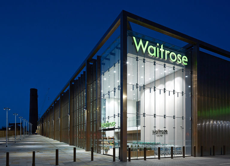 Evening photo of exterior façade to Waitrose in Chester, with full height glazing and bronze metal cladding elements.